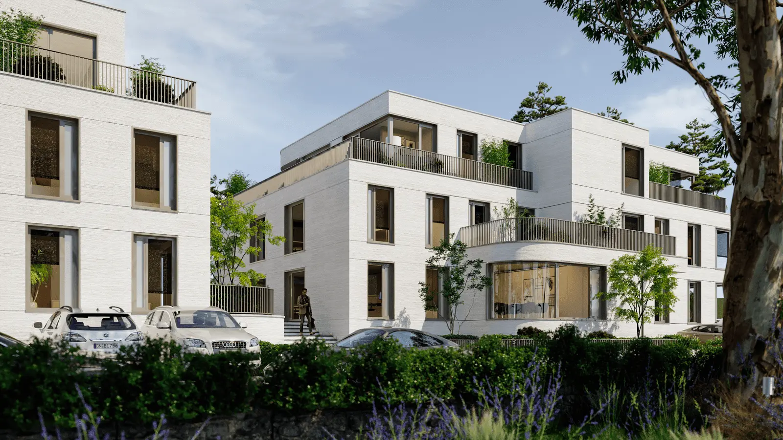 Modernes, weißes, dreistöckiges Wohnhaus mit großen Fenstern, Balkonen und Grünanlagen; davor sind mehrere geparkte Autos zu sehen.