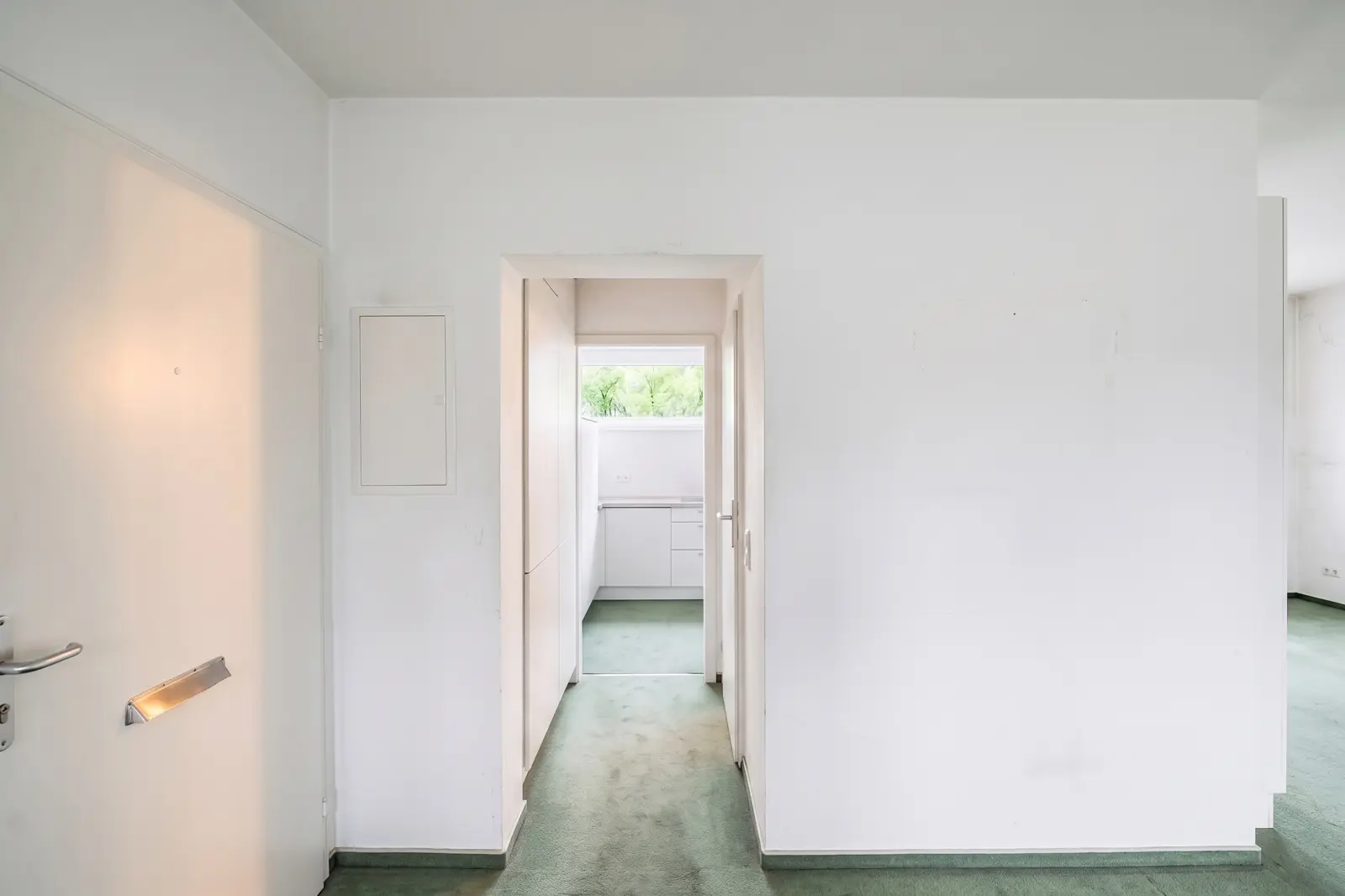 Blick auf einen weißen Flur mit hellgrünem Teppichboden, der zu einer kleinen Küche mit einem Fenster am anderen Ende führt. Der Flur hat schlichte Wände und ist nur minimal dekoriert.
