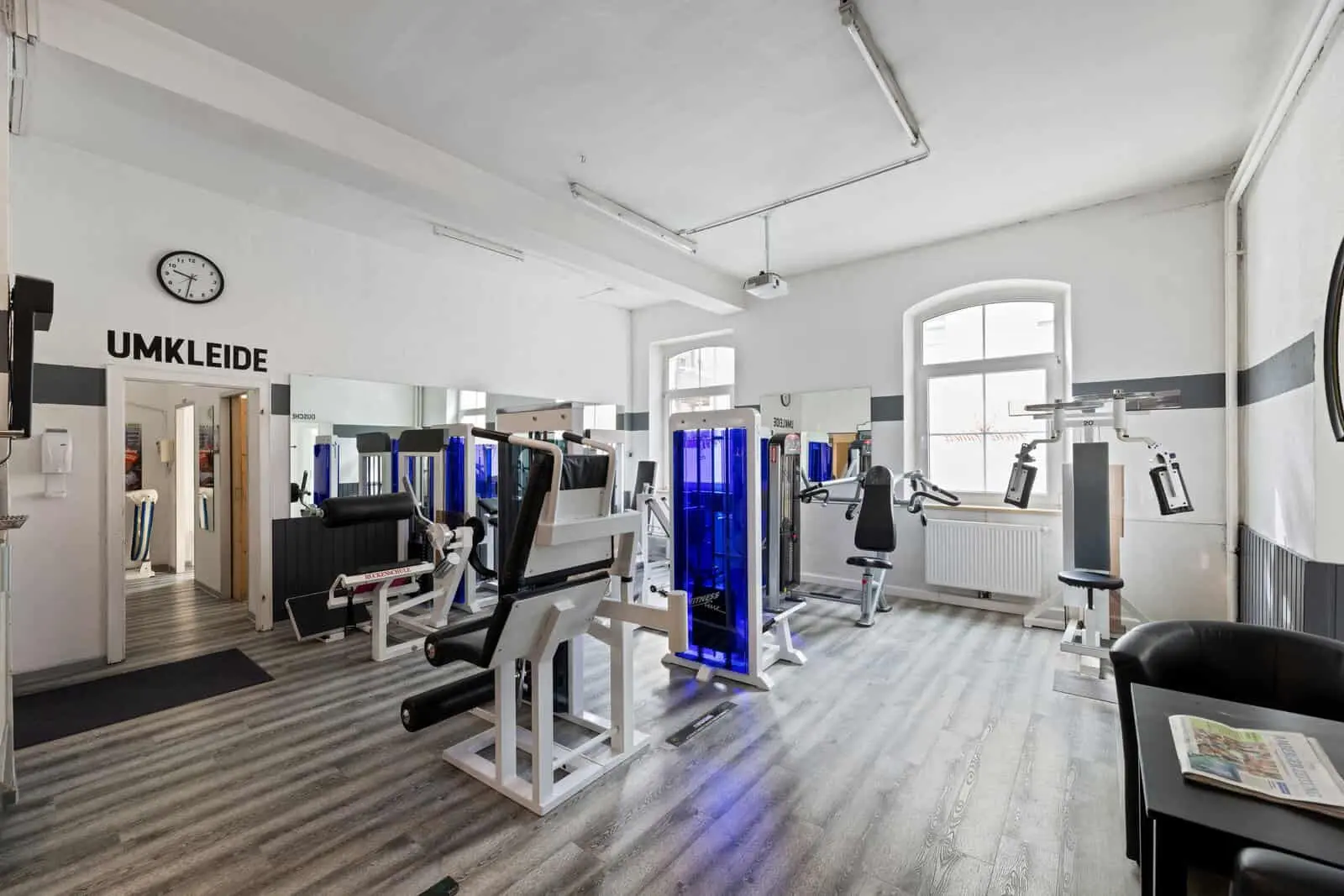 Ein Fitnessraum mit verschiedenen Kraftgeräten, Spiegeln an den Wänden, einer Uhr über einer Tür mit der Aufschrift "Umkleide" und einem Stuhl mit Zeitungen auf einem Beistelltisch.