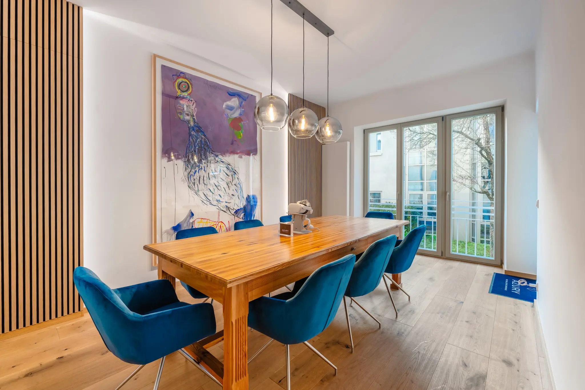 Das Esszimmer verfügt über einen Holztisch, sechs blaue Stühle und drei kugelförmige Hängelampen. Ein großes Pfauen-Kunstwerk ziert die Wand. Glastüren führen auf einen gemütlichen Balkon mit Blick auf Bäume – eine charmante „Über uns“-Note, die dem Raum Charakter verleiht.