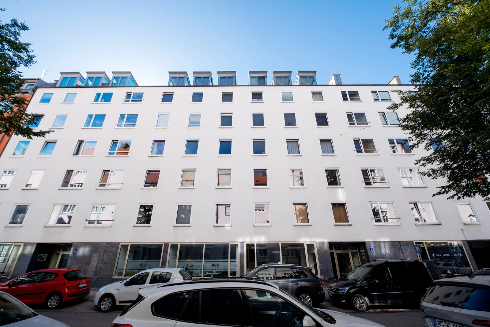 Ein fünfstöckiges weißes Wohnhaus mit unregelmäßig angeordneten Fenstern und geparkten Autos entlang der Straße davor.