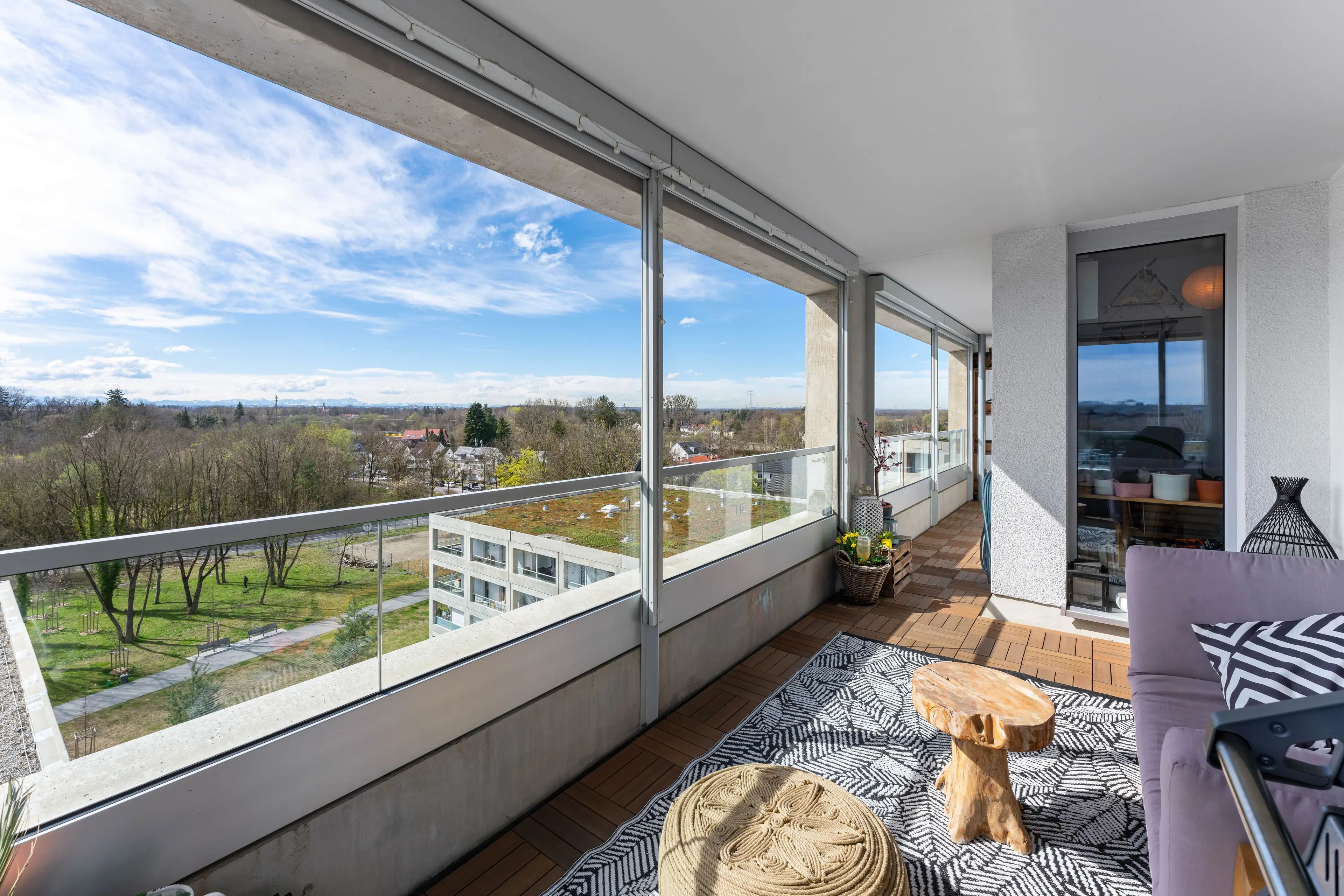 Ein geräumiger Balkon mit Holzboden und gemustertem Teppich bietet Ausblicke auf einen Park und die Stadtlandschaft. Glasgeländer unterstreichen das moderne Design. Ein Tisch und dekorative Gegenstände sind ordentlich platziert.