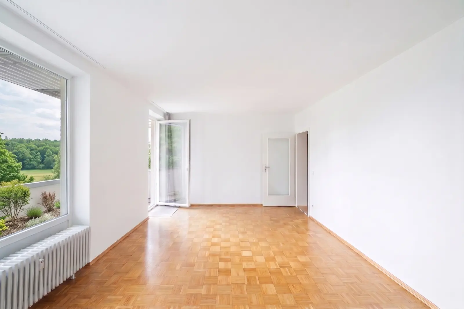 Ein leerer Raum mit weißen Wänden, Parkettboden, einem großen Fenster, einer Glastür zum Balkon und einem Heizkörper unter dem Fenster.