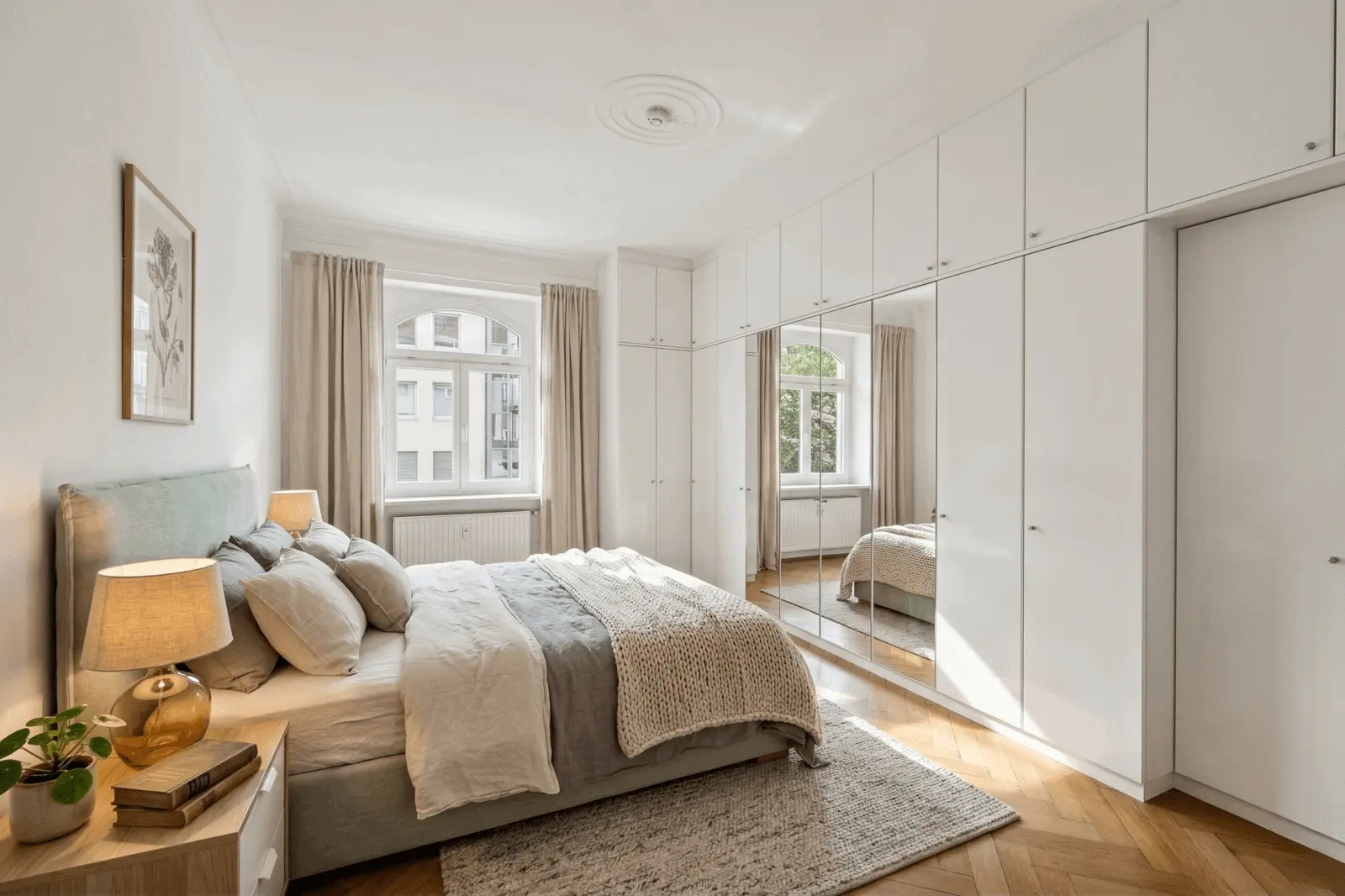 Ein modernes Schlafzimmer mit einem Doppelbett, Nachttischen, einer Lampe, einem großen Kleiderschrank mit Spiegeln, hellen Vorhängen und einem Fenster, das Tageslicht hereinlässt.