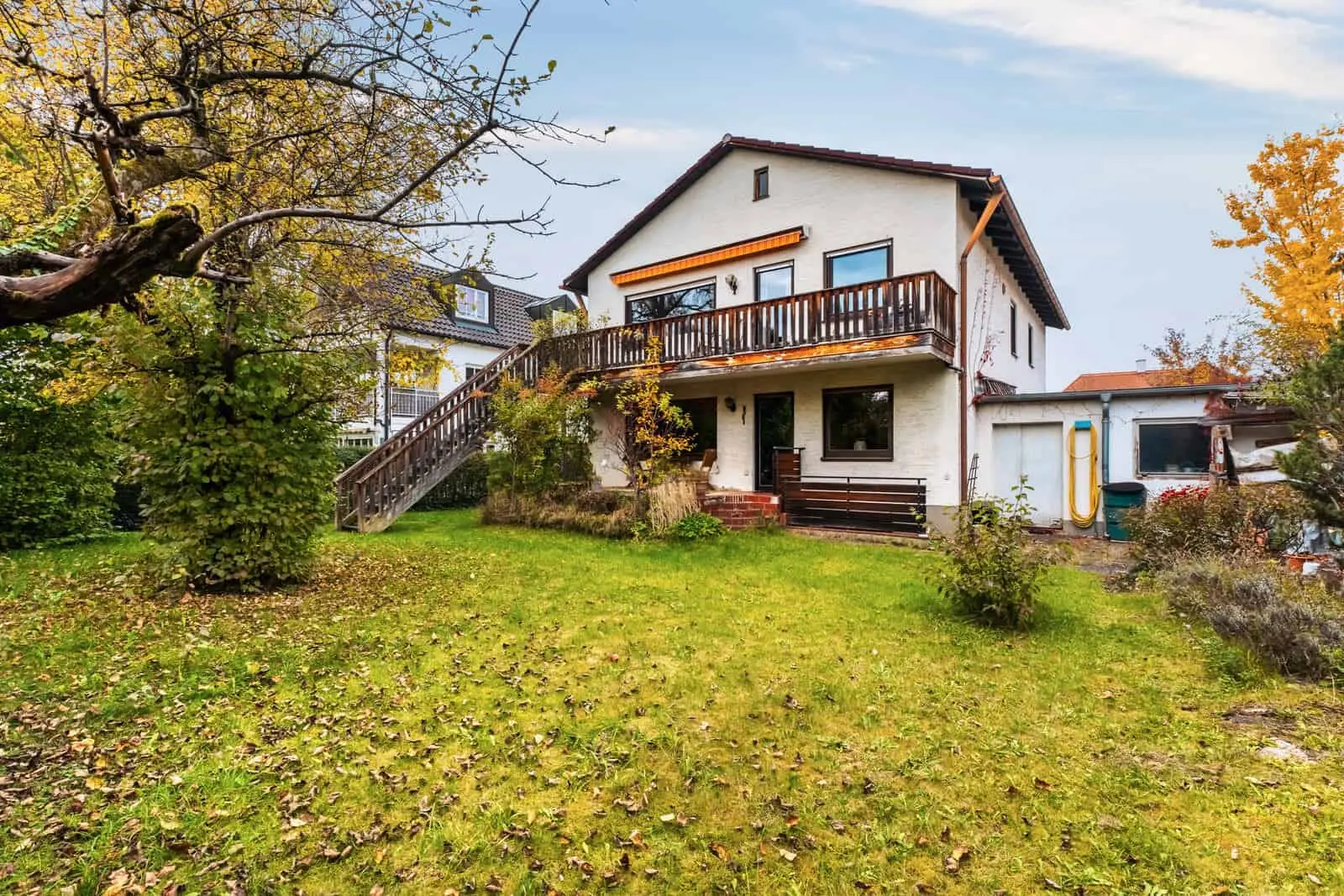 Ein zweistöckiges weißes Haus mit einem Holzbalkon und einer Außentreppe, umgeben von einem grasbewachsenen Hof mit Bäumen und Herbstlaub.
