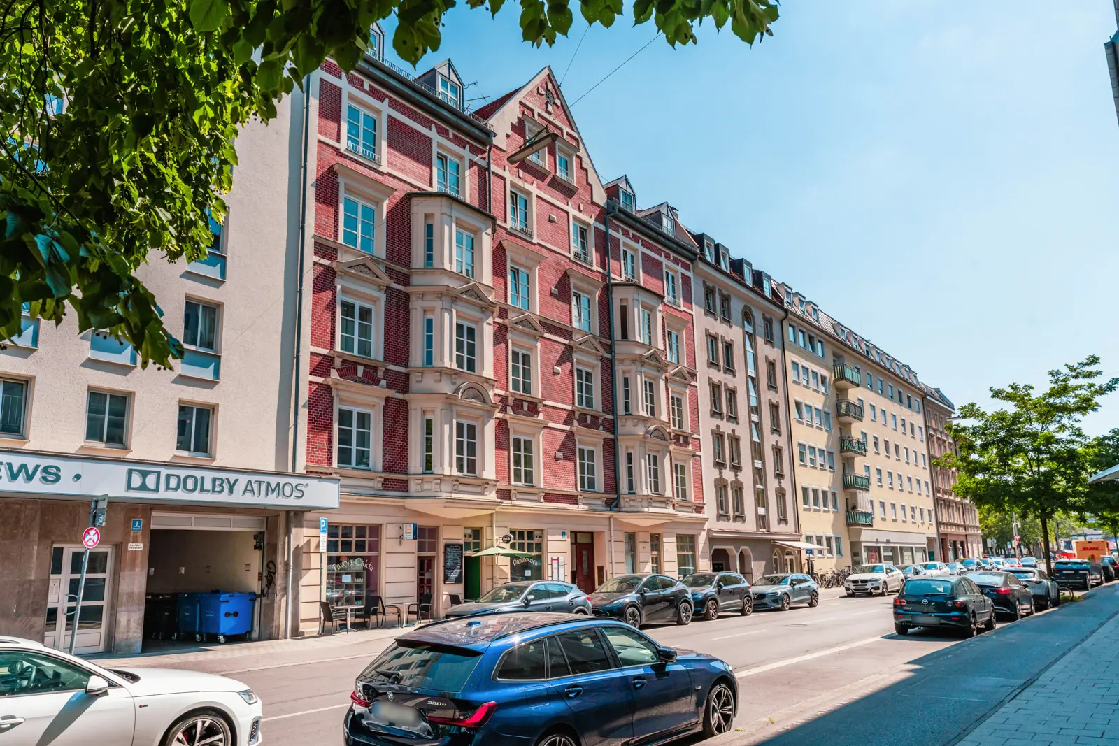 Eine Stadtstraße mit geparkten Autos, die an einem sonnigen Tag vor mehrstöckigen Wohn- und Geschäftsgebäuden reihenweise stehen.