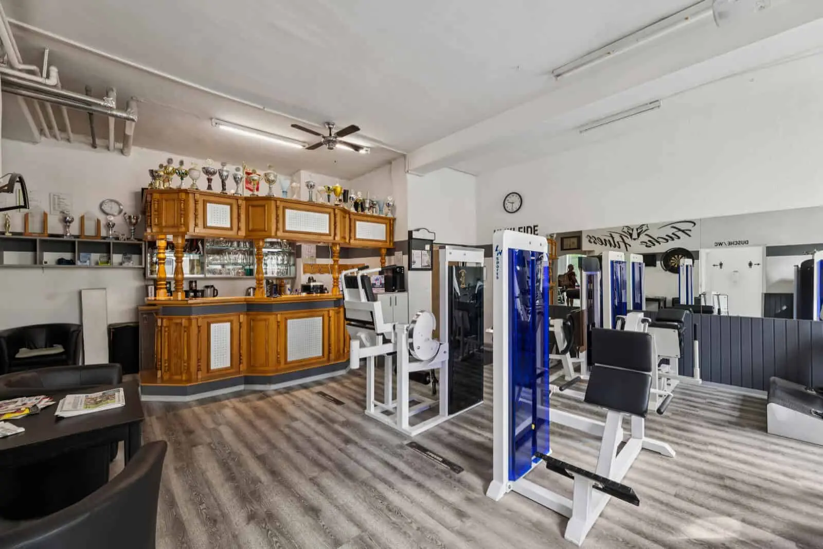Ein Fitnessstudio mit Fitnessgeräten im Vordergrund und einer hölzernen Theke mit Trophäen und Spiegeln im Hintergrund.
