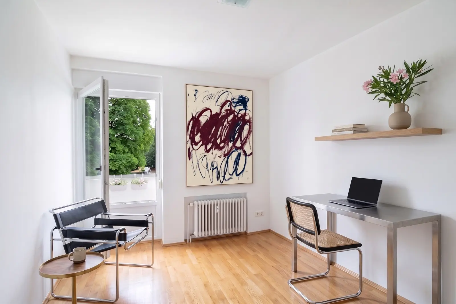 Minimalistisches Homeoffice mit einem Schreibtisch, einem Laptop, einem Stuhl, zwei Sesseln, einer Kaffeetasse, abstrakter Wandkunst, einem schwebenden Regal mit Büchern und Blumen sowie einer offenen Tür, die auf einen Balkon führt.