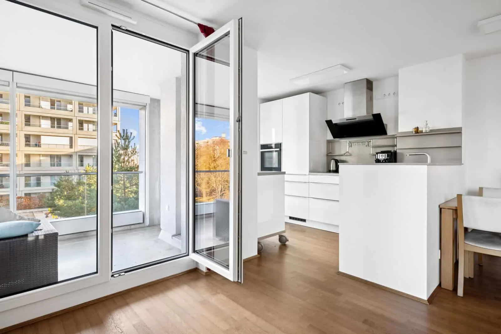 Moderne weiße Küche mit Holzboden, offenen Glastüren, die auf einen Balkon führen, und einem Sitzbereich im Freien mit Blick auf Wohnhäuser und Bäume.