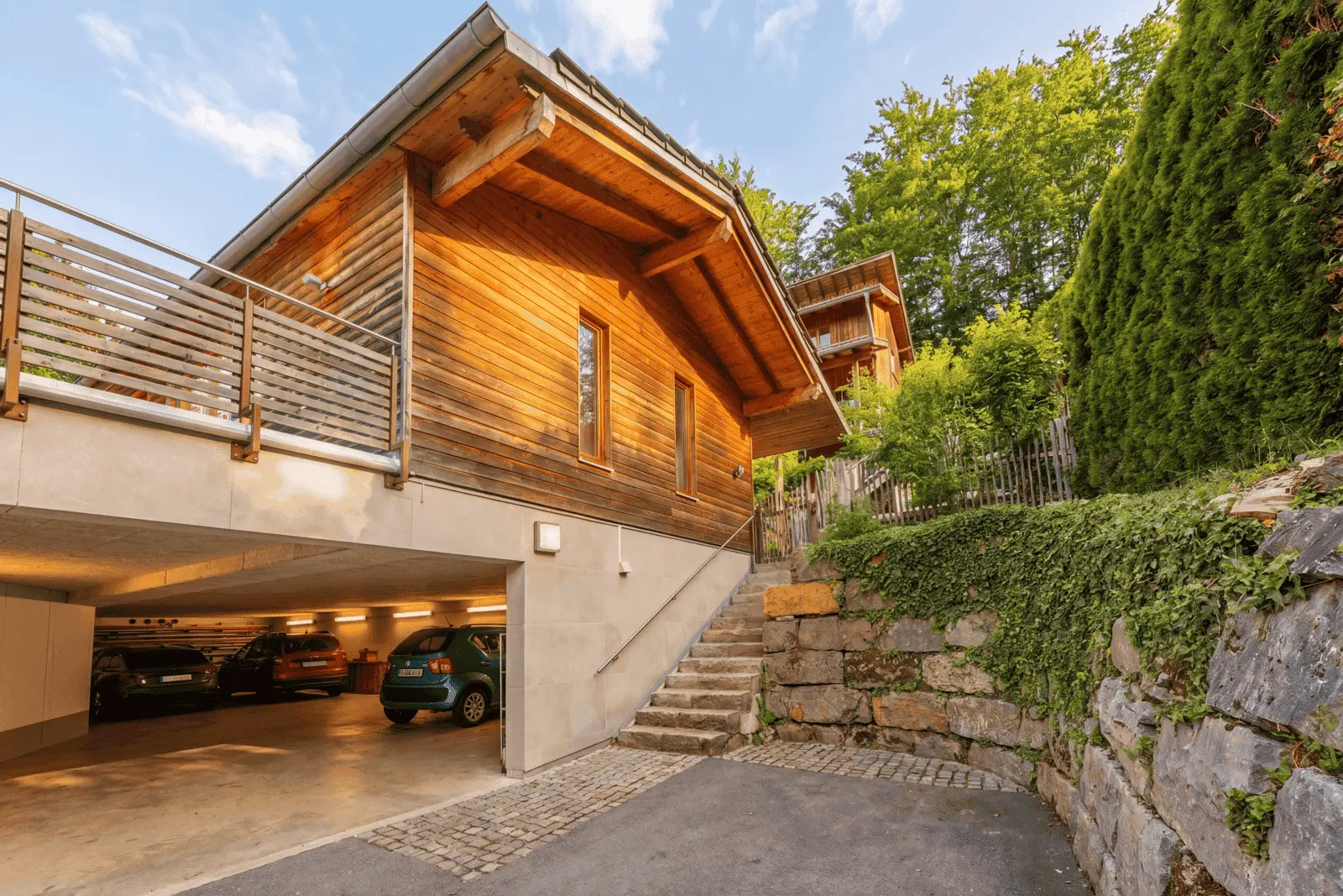 Ein modernes Holzhaus mit einer offenen Garage darunter, geparkten Autos, einer Steintreppe, die hinaufführt, und Grünflächen rund um das Grundstück.