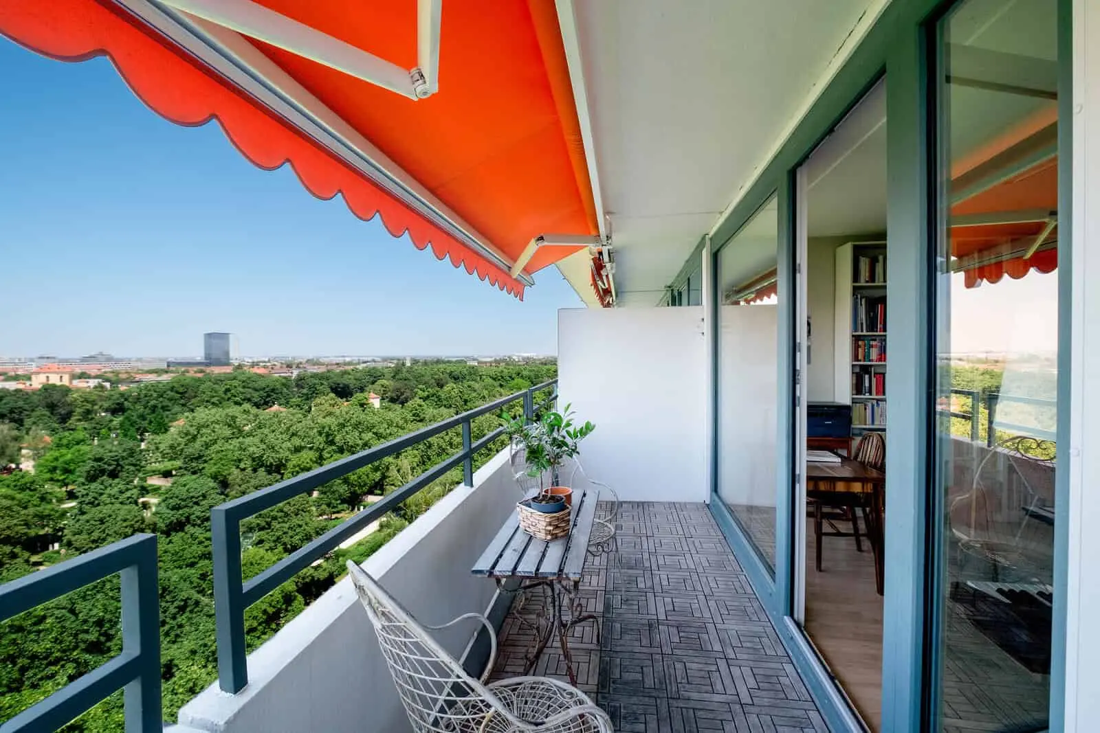 Ein Balkon mit Tisch, Stuhl und orangefarbener Markise bietet einen Blick auf einen grünen Park und die Stadt, und Glastüren führen in einen Raum mit Bücherregalen und einem Esstisch aus Holz.