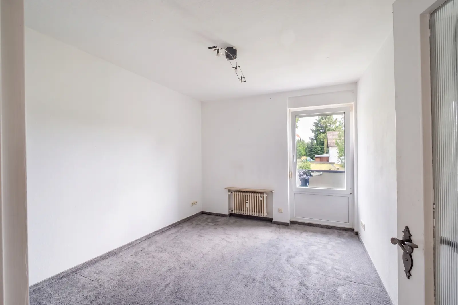 Ein leerer Raum mit weißen Wänden, grauem Teppichboden, einem Fenster, durch das Tageslicht hereinfällt, einem Heizkörper unter dem Fenster und einer auf der rechten Seite teilweise geöffneten Tür. Es ist eine Deckenleuchte vorhanden.