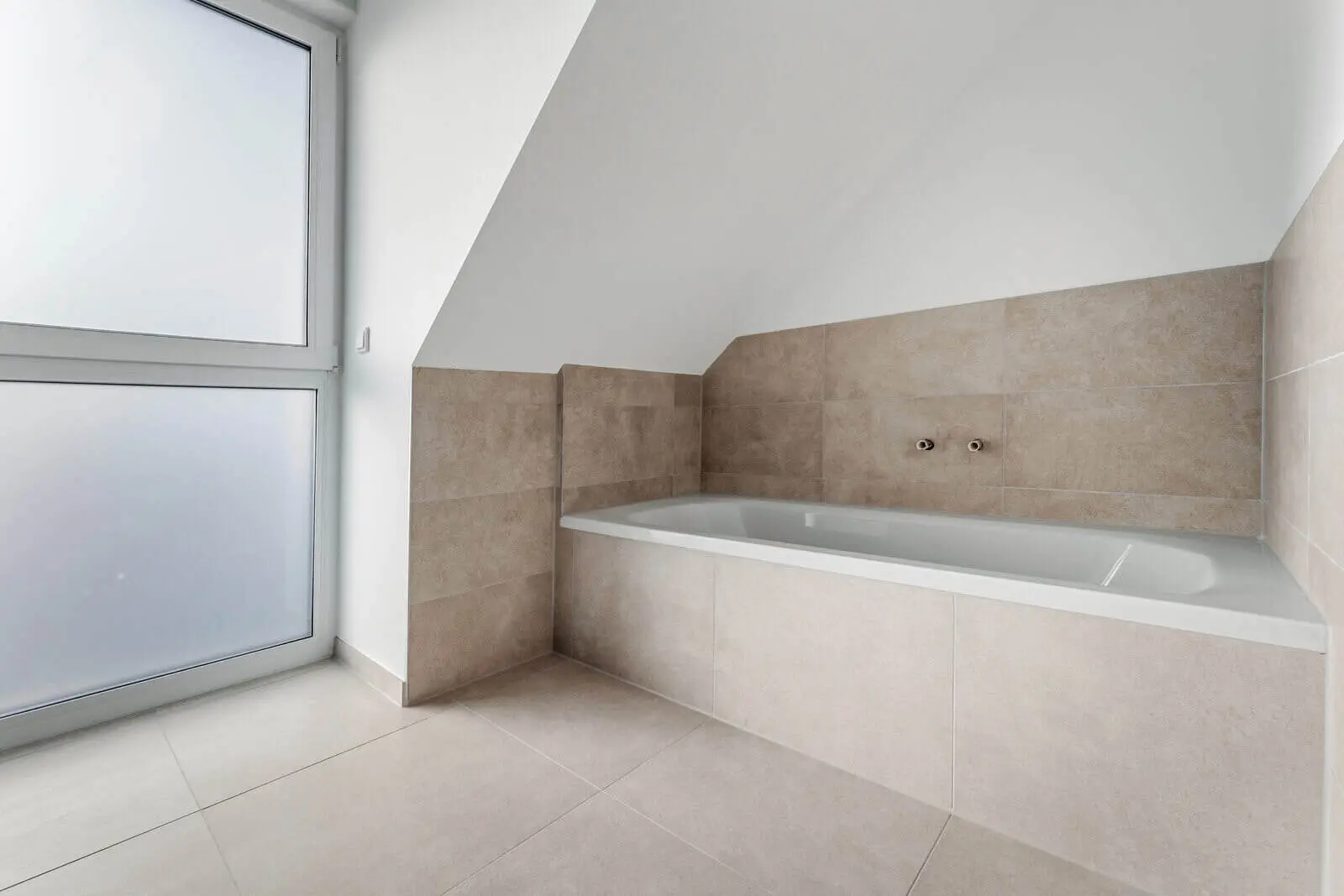 Modernes Badezimmer mit eingebauter Badewanne, beige gefliesten Wänden und Böden sowie einem großen Milchglasfenster, das für Tageslicht sorgt.