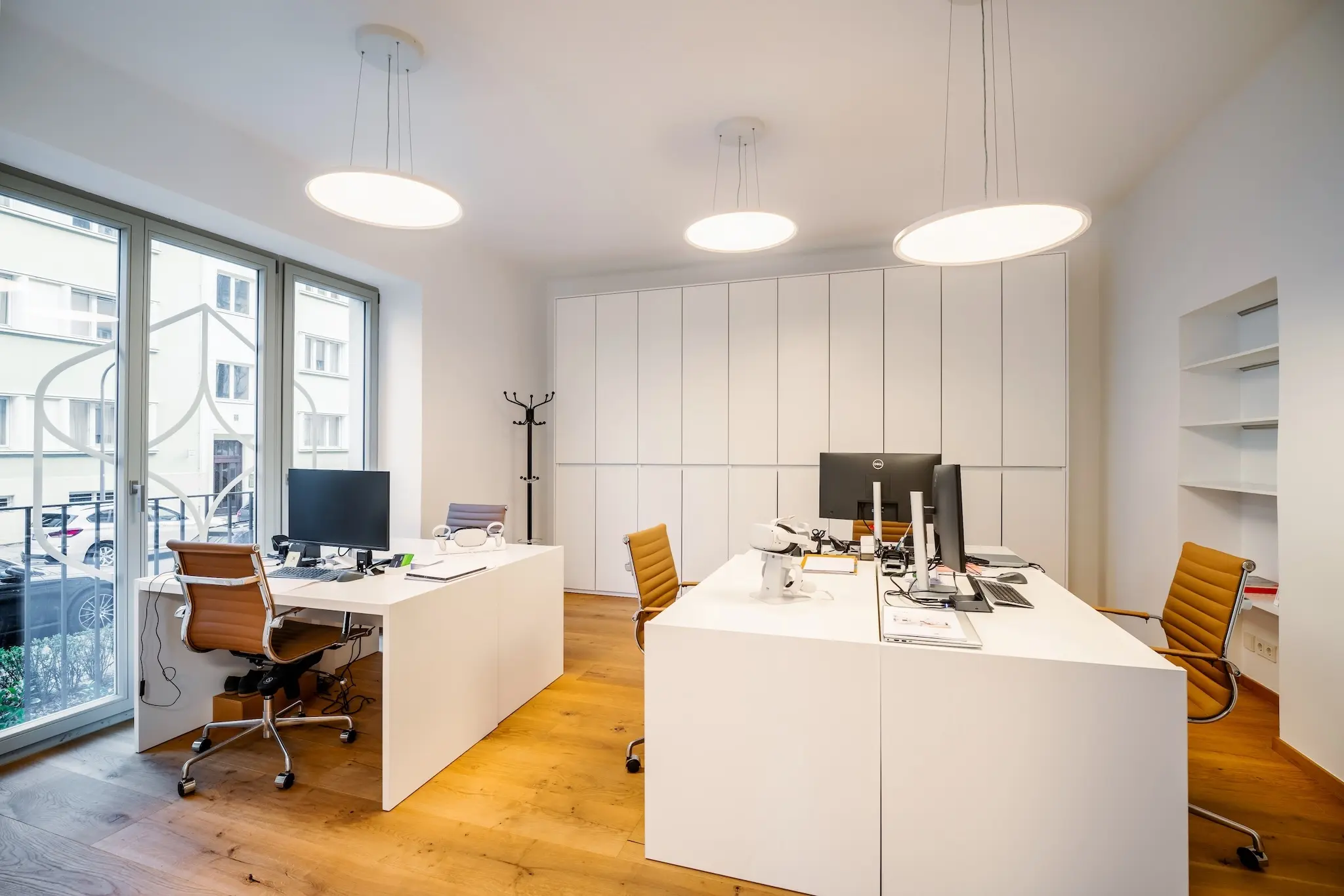 Ein modernes Büro mit zwei Holzschreibtischen, braunen Stühlen und Computern. Deckenlampen erhellen den Raum, der Holzböden und weiße Wände mit großen Fenstern auf der linken Seite aufweist.