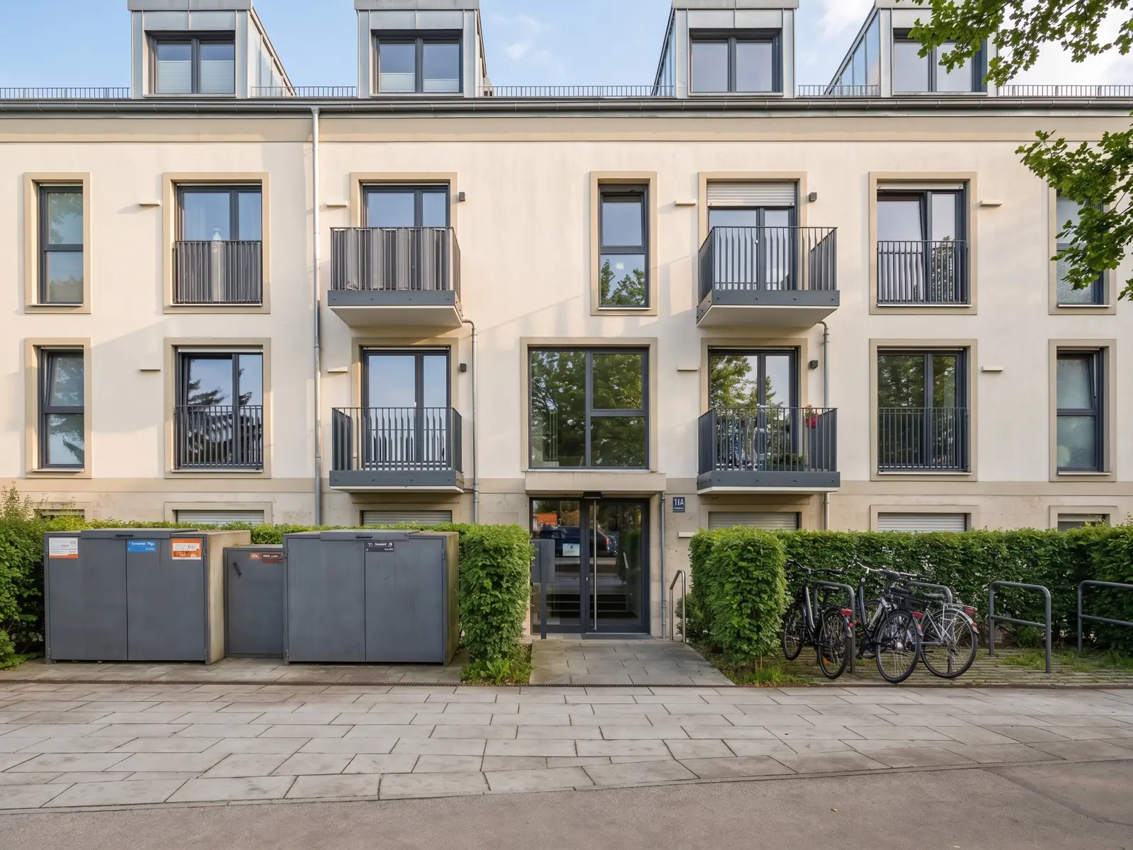 Modernes dreistöckiges Mehrfamilienhaus mit Balkonen, einem verglasten Eingangsbereich und vor dem Haus abgestellten Fahrrädern. Mülltonnen und Hecken säumen den Bürgersteig.