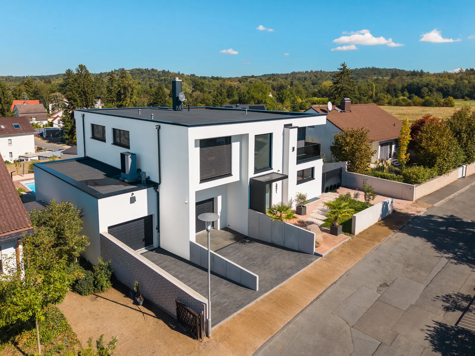 Luftaufnahme eines zweistöckigen, modernen, weißen Hauses mit Flachdach, Garage und gepflasterter Einfahrt in einem Vorortviertel.