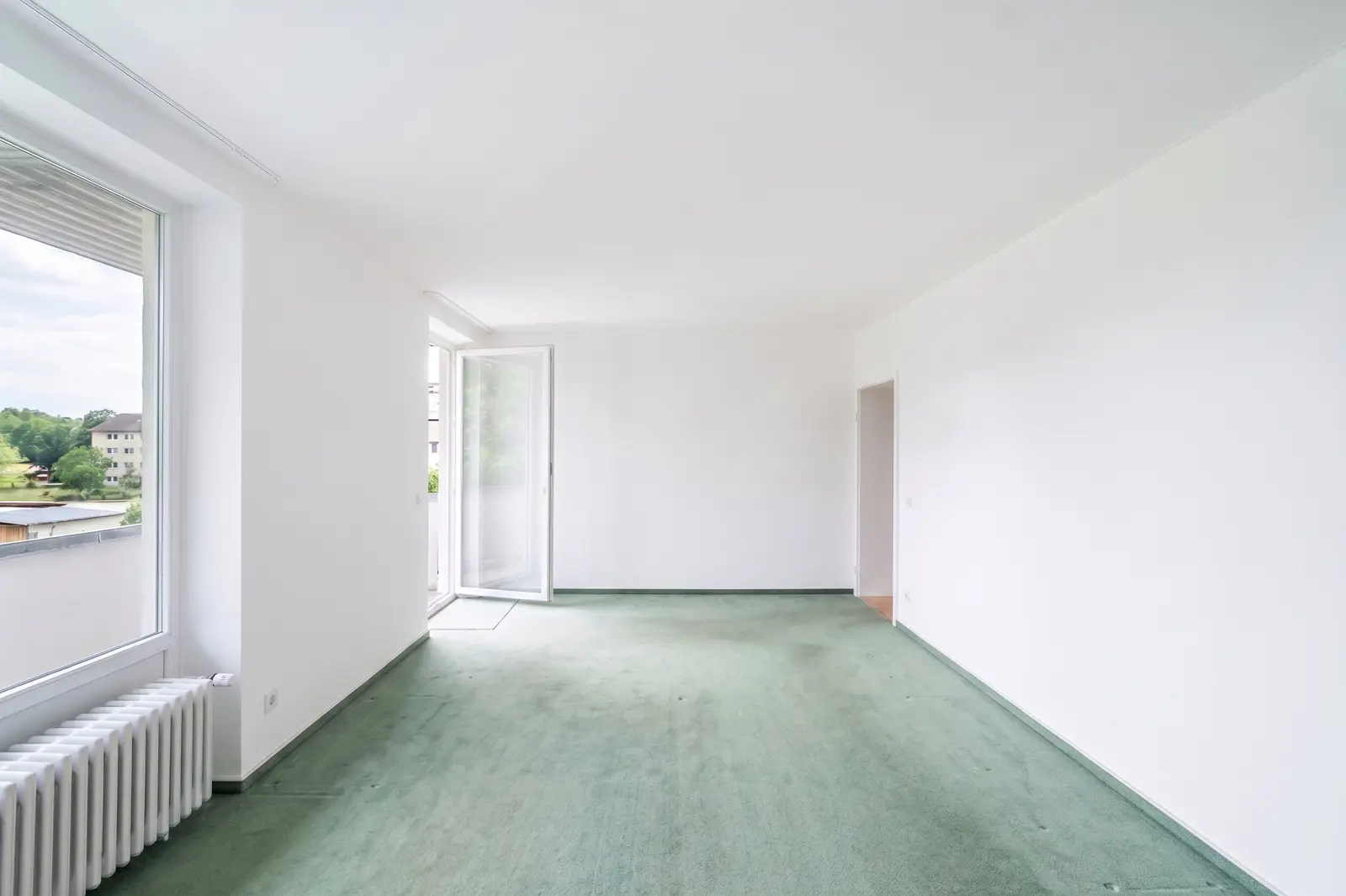 Ein leerer Raum mit weißen Wänden, grünem Teppichboden, einem Heizkörper unter einem Fenster, einer offenen Tür zum Balkon und einer Tür zu einem anderen Raum.
