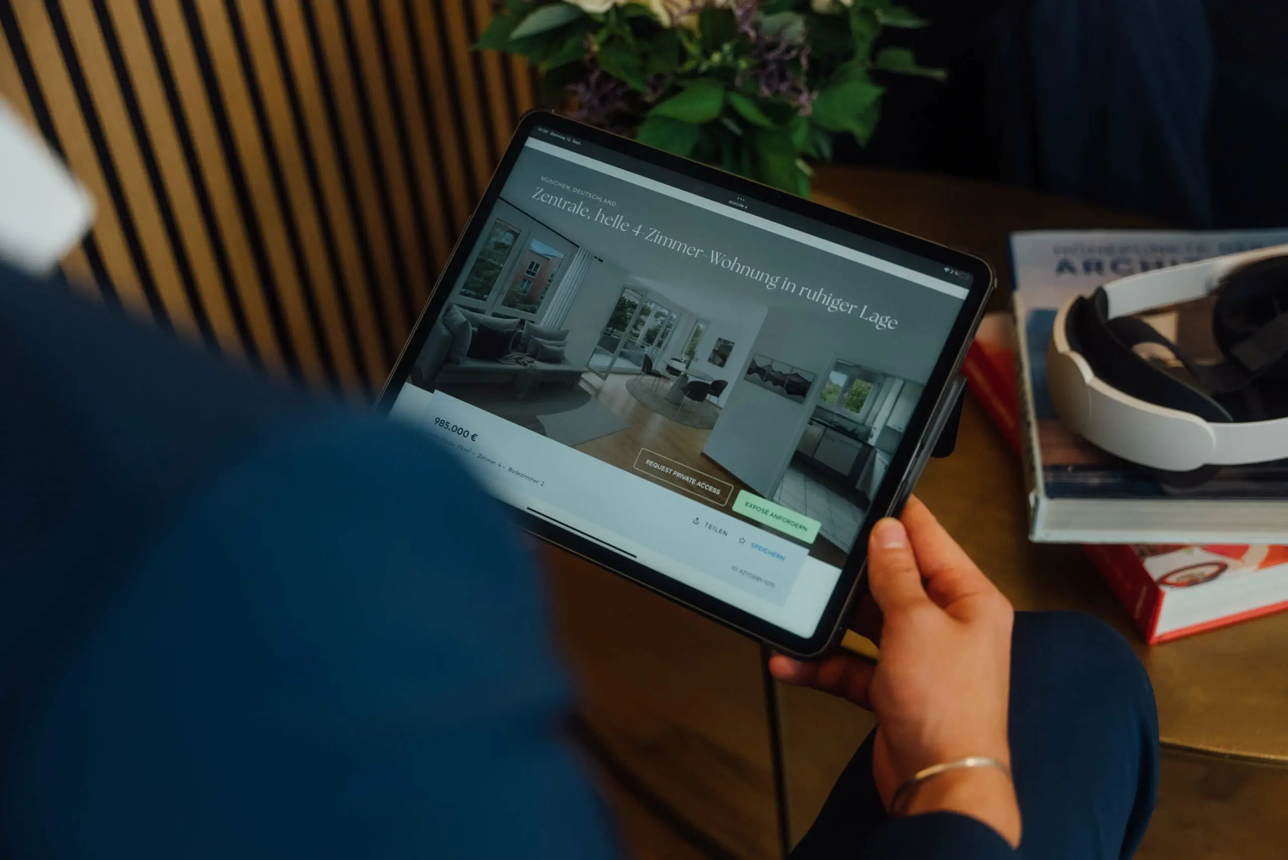 Eine Person hält ein Tablet, auf dem eine Immobilien-Website mit atemberaubenden Bildern eines Wohnzimmers präsentiert wird. Daneben liegen ein VR-Headset und Bücher auf dem Tisch und deuten damit die immersive Welt an, in der Technologie auf den Hauskauf trifft. Erfahren Sie mehr über uns in dieser nahtlosen Mischung aus Innovation und Komfort.