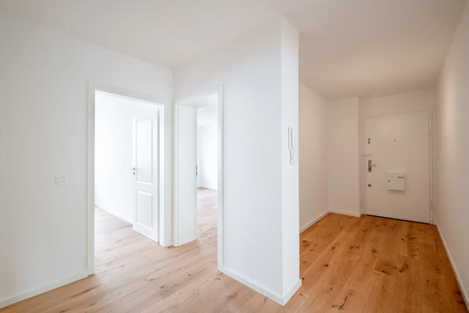 Ein heller, leerer Flur in einer Wohnung mit hellem Holzboden, weißen Wänden und Türen, die zu anderen Räumen führen. Am Ende des Flurs ist die Haustür zu sehen.