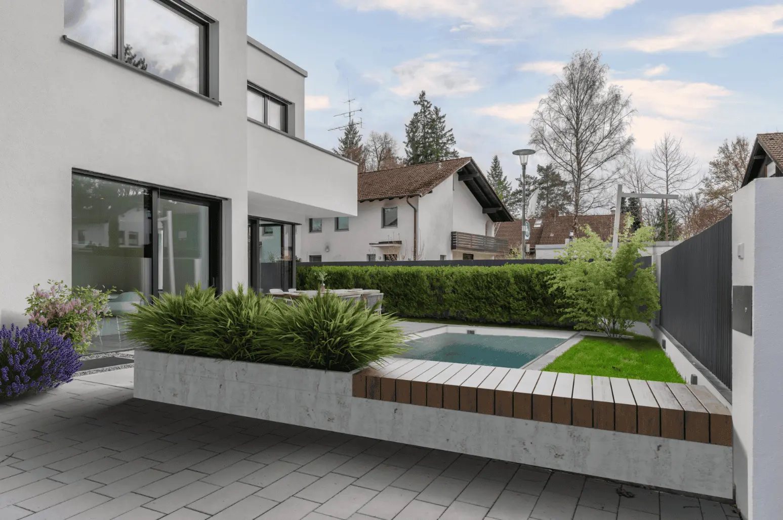 Modernes Haus mit großen Fenstern, einem begrünten Hof, einem rechteckigen Pool, einer Holzbank und benachbarten Häusern im Hintergrund unter einem teilweise bewölkten Himmel.