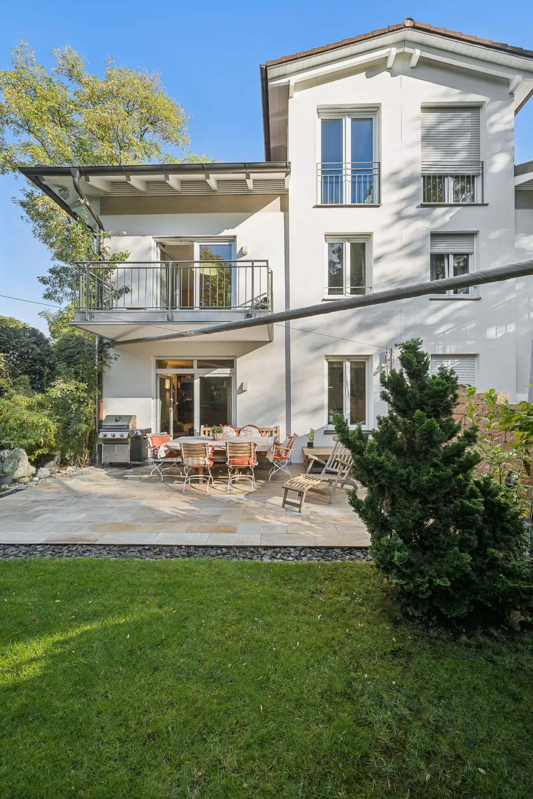Zweigeschossiges weißes Haus mit einer Terrasse aus Stein, einer Essgruppe im Freien, einem Grill und einem Rasen.