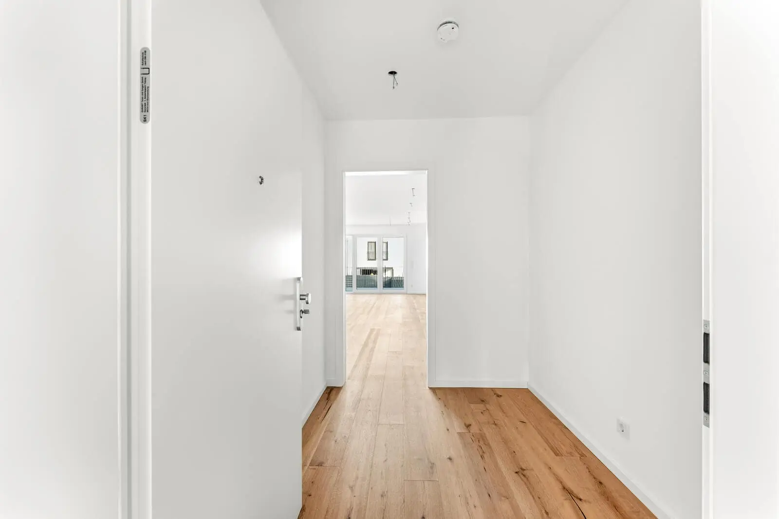 Ein heller, leerer Flur mit weißen Wänden und Holzboden führt zu einem geräumigen Zimmer mit großen Fenstern im Hintergrund.