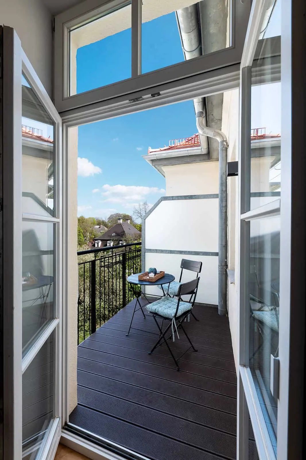 Kleiner Balkon mit schwarzem Metallgeländer, zwei Stühlen und einem kleinen Tisch mit einem Tablett, der an einem sonnigen Tag durch eine offene Doppelglastür zu sehen ist.