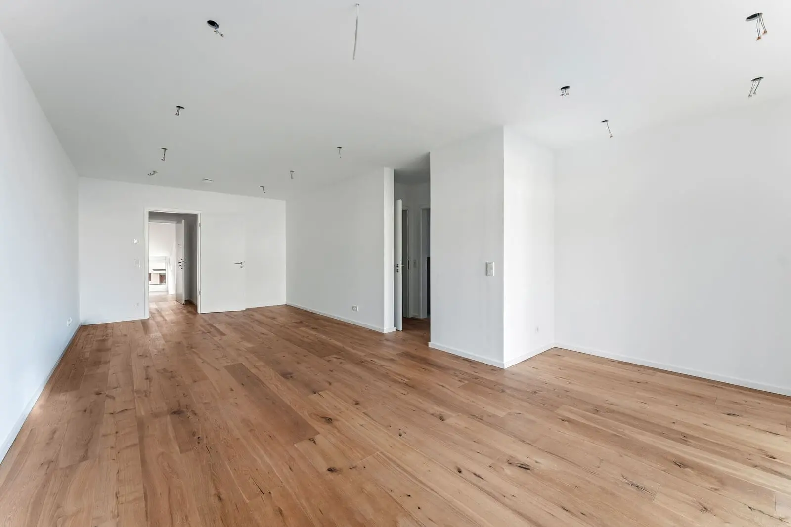 Leerer Raum mit weißen Wänden, Holzboden, sichtbaren Deckenleuchten und offenen Türen, die zu anderen Räumen führen.