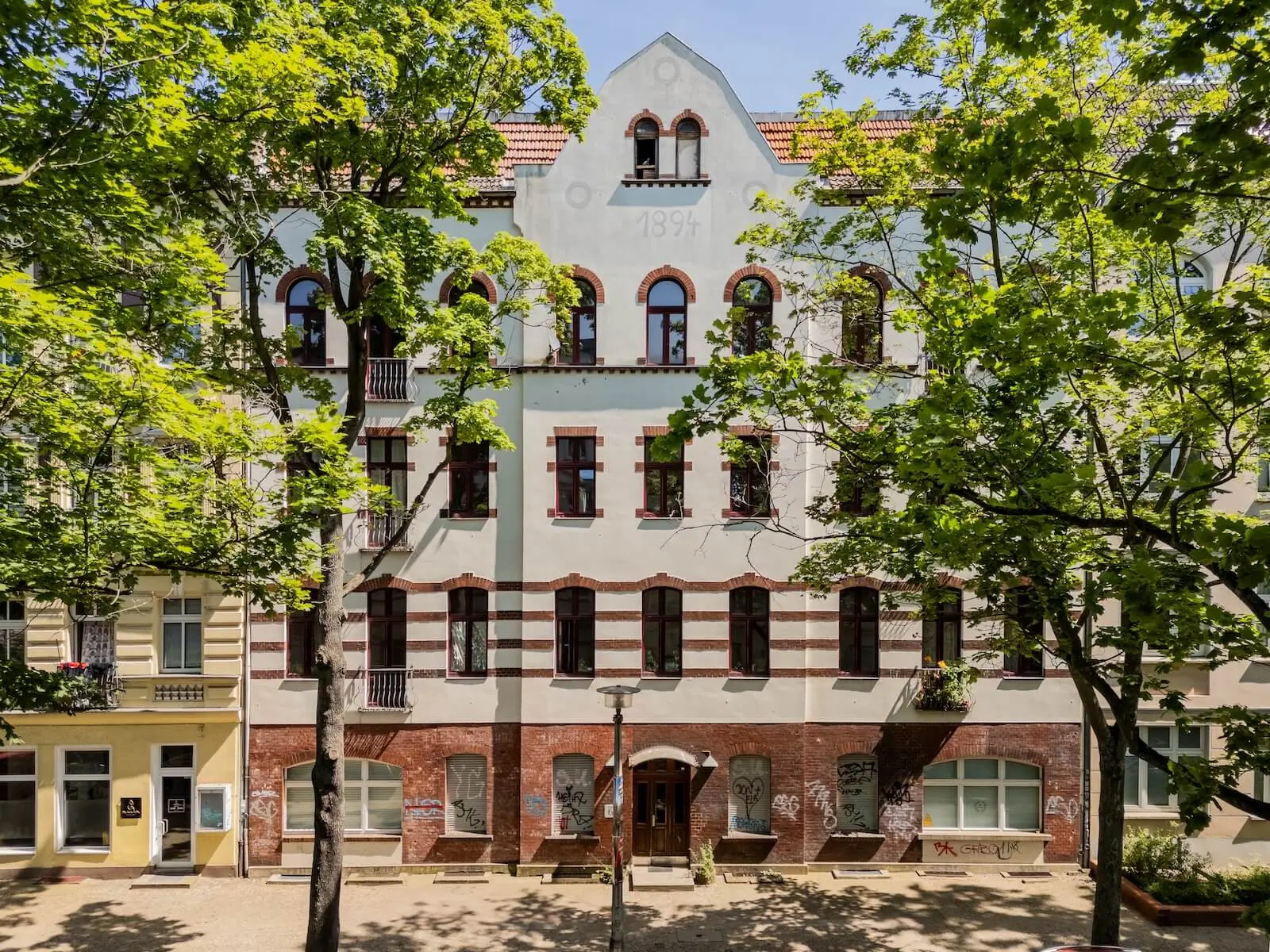 Ein mehrstöckiges Mehrfamilienhaus in Berlin mit Bogenfenstern und Graffiti an der unteren Fassade, umgeben von grünen Bäumen.
