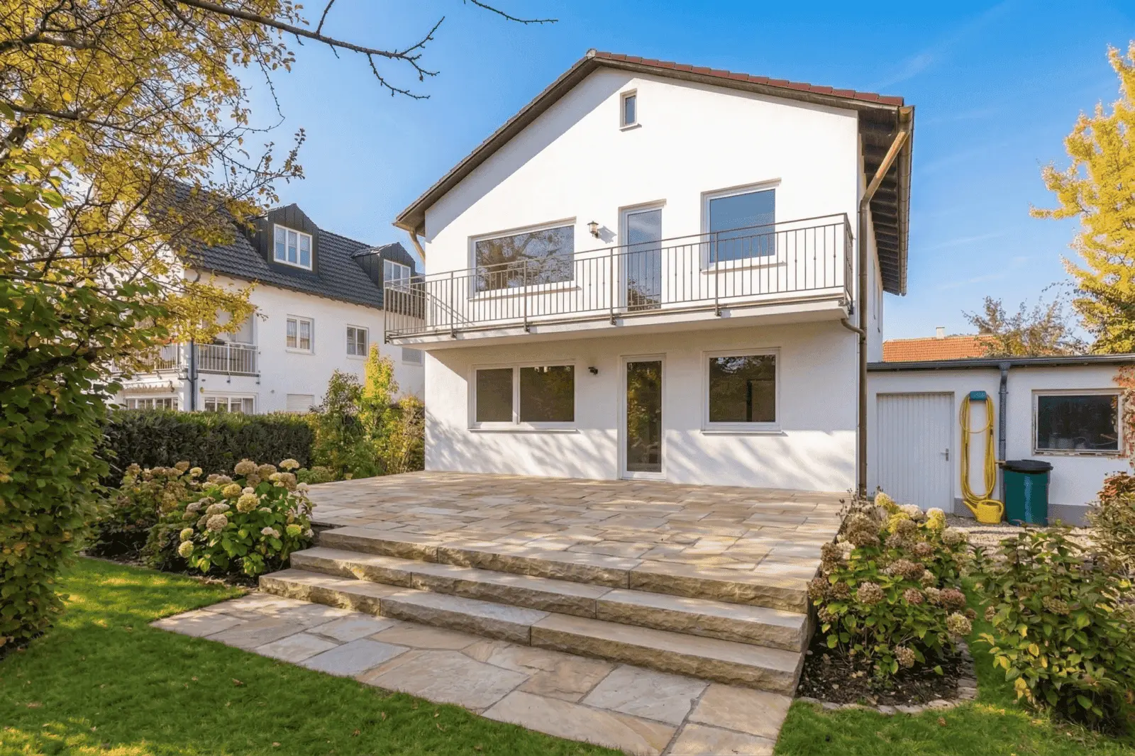 Zweistöckiges weißes Haus mit einem Balkon, großen Fenstern, einer Steinterrasse und einer Treppe, die zu einem grünen Rasen führt, umgeben von Sträuchern und Nachbarhäusern.