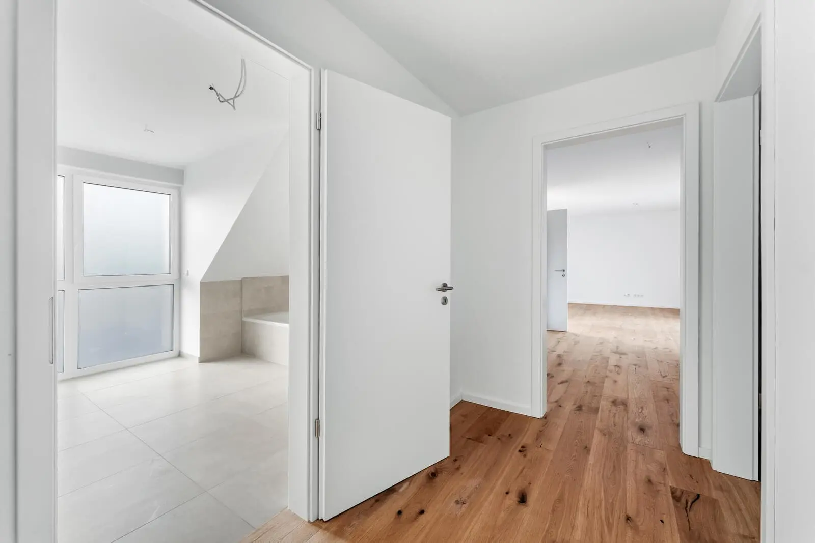 Über einen Flur mit Holzboden gelangt man in ein gefliestes Bad mit Badewanne und einen hellen, leeren Raum mit großen Fenstern. Die Wände und Türen sind weiß.