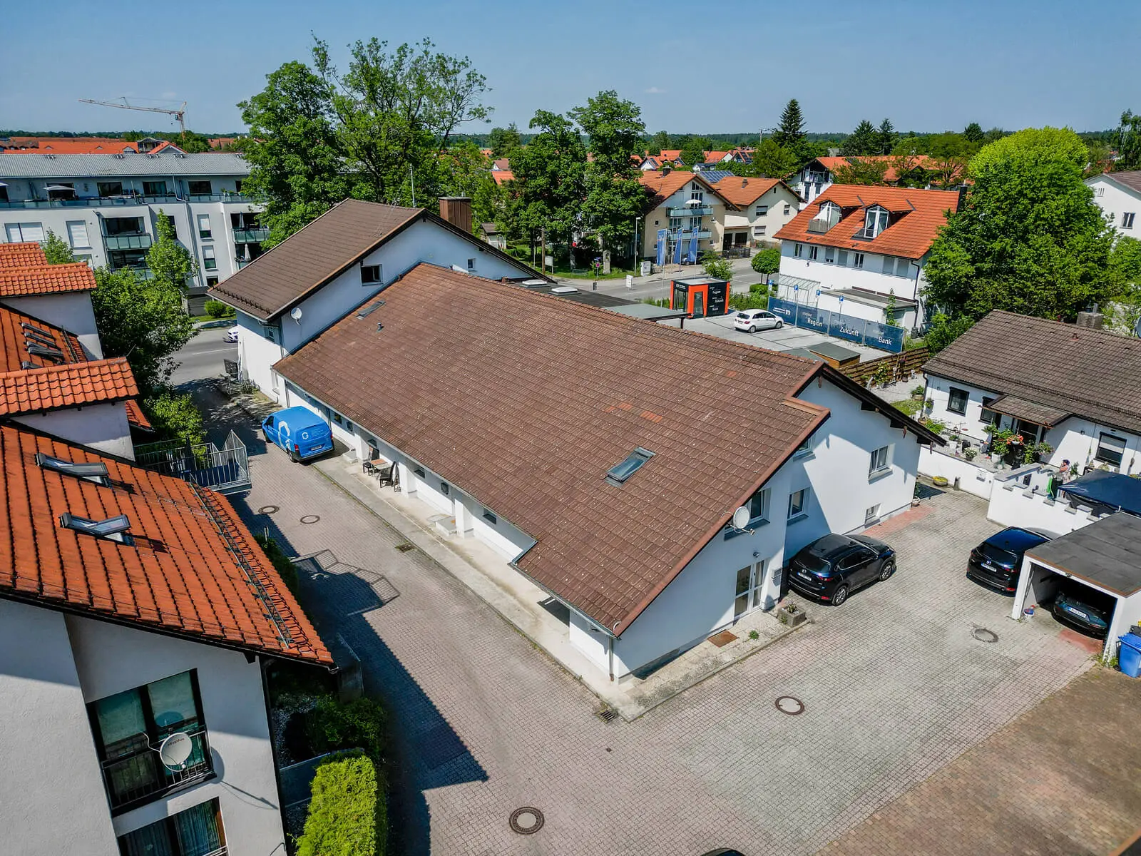 Eine Luftaufnahme von Wohnhäusern mit roten Ziegeldächern und umgebendem Grün an einem sonnigen Tag. In der Nähe der Gebäude sind mehrere geparkte Autos zu sehen.