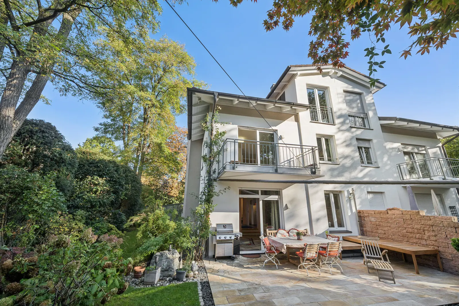 Ein weißes, zweistöckiges Haus mit einer Terrasse auf der Rückseite, auf der eine Essgruppe, ein Grill und eine Steinmauer stehen.