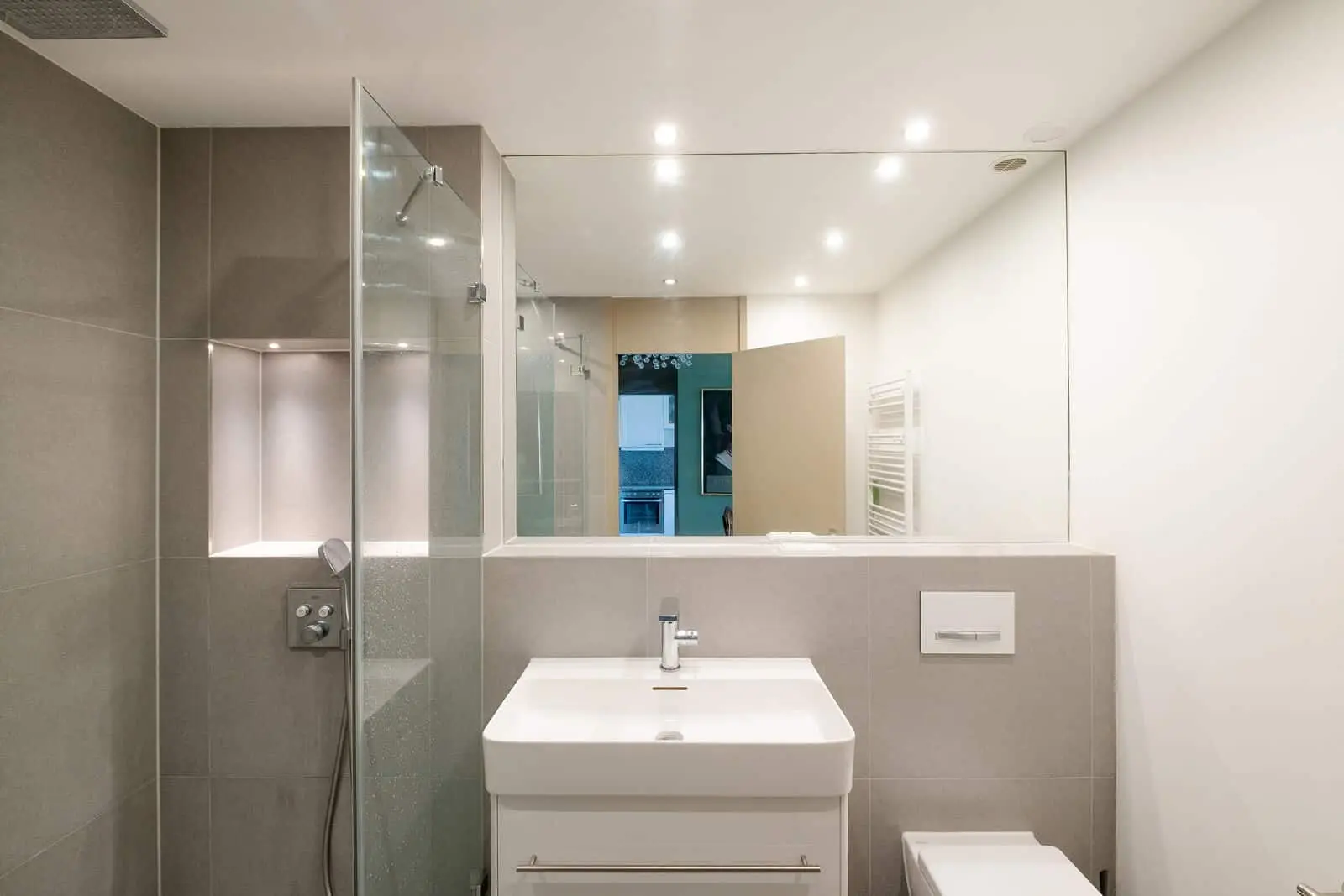 Modernes Badezimmer mit begehbarer Glasdusche, großem Wandspiegel, weißem Waschbecken mit Unterschrank, wandmontierter Toilette und neutralem Dekor in Grau und Weiß.