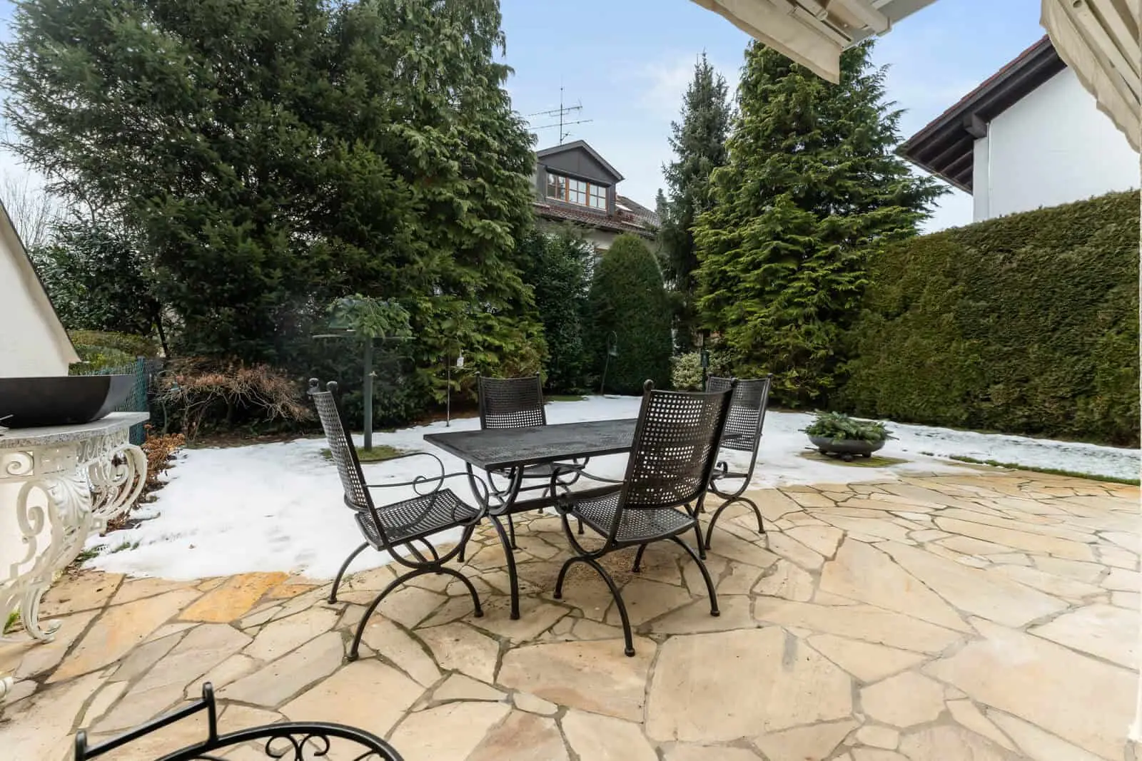 Terrassenbereich mit schwarzem Metalltisch und vier Stühlen auf Steinboden, umgeben von Schneeflecken, immergrünen Bäumen und Hecken.