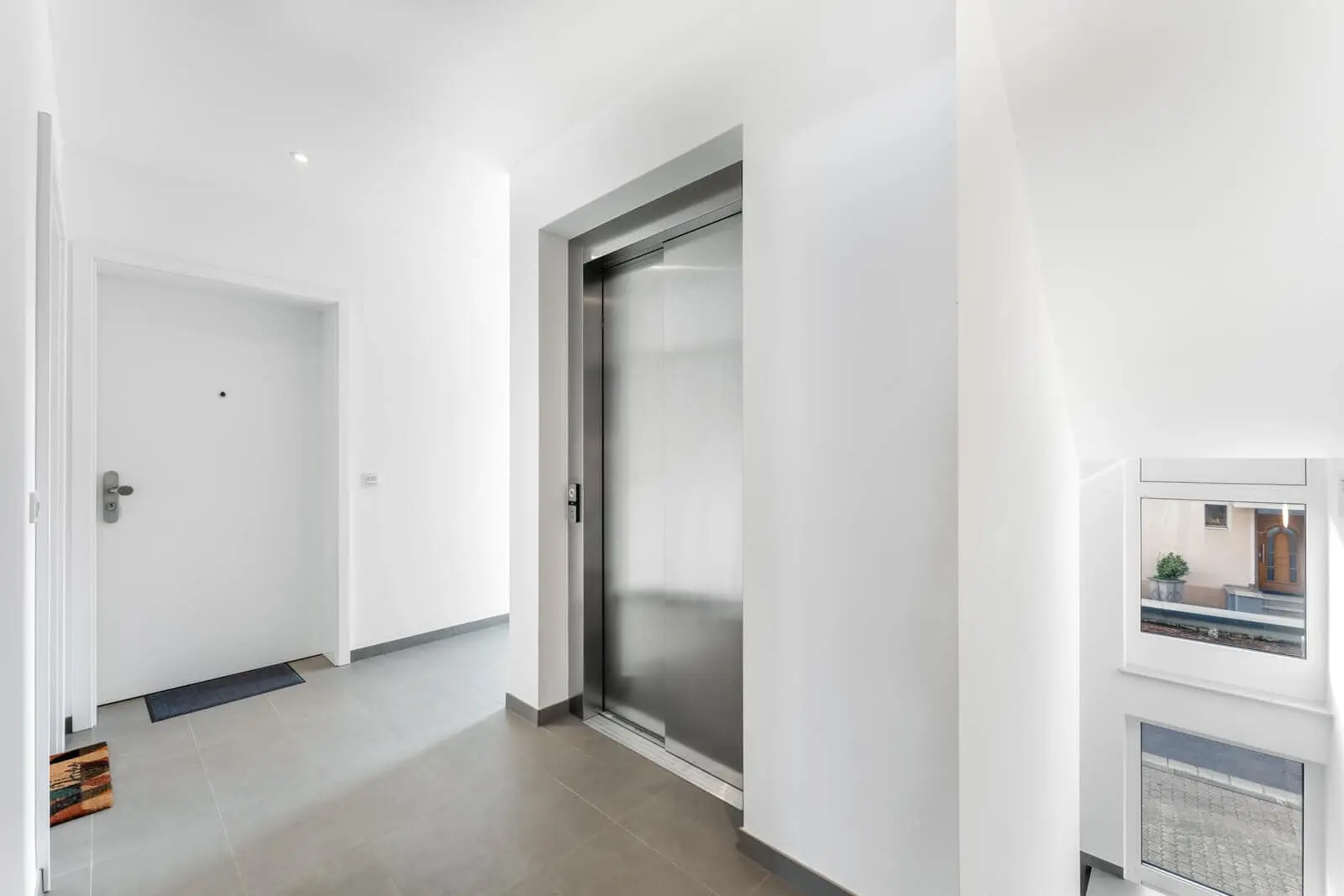 Moderner Wohnhausflur mit geschlossener Aufzugstür, weißer Eingangstür, hellgrauem Fliesenboden und einem Fenster in der Nähe eines Treppenhauses.