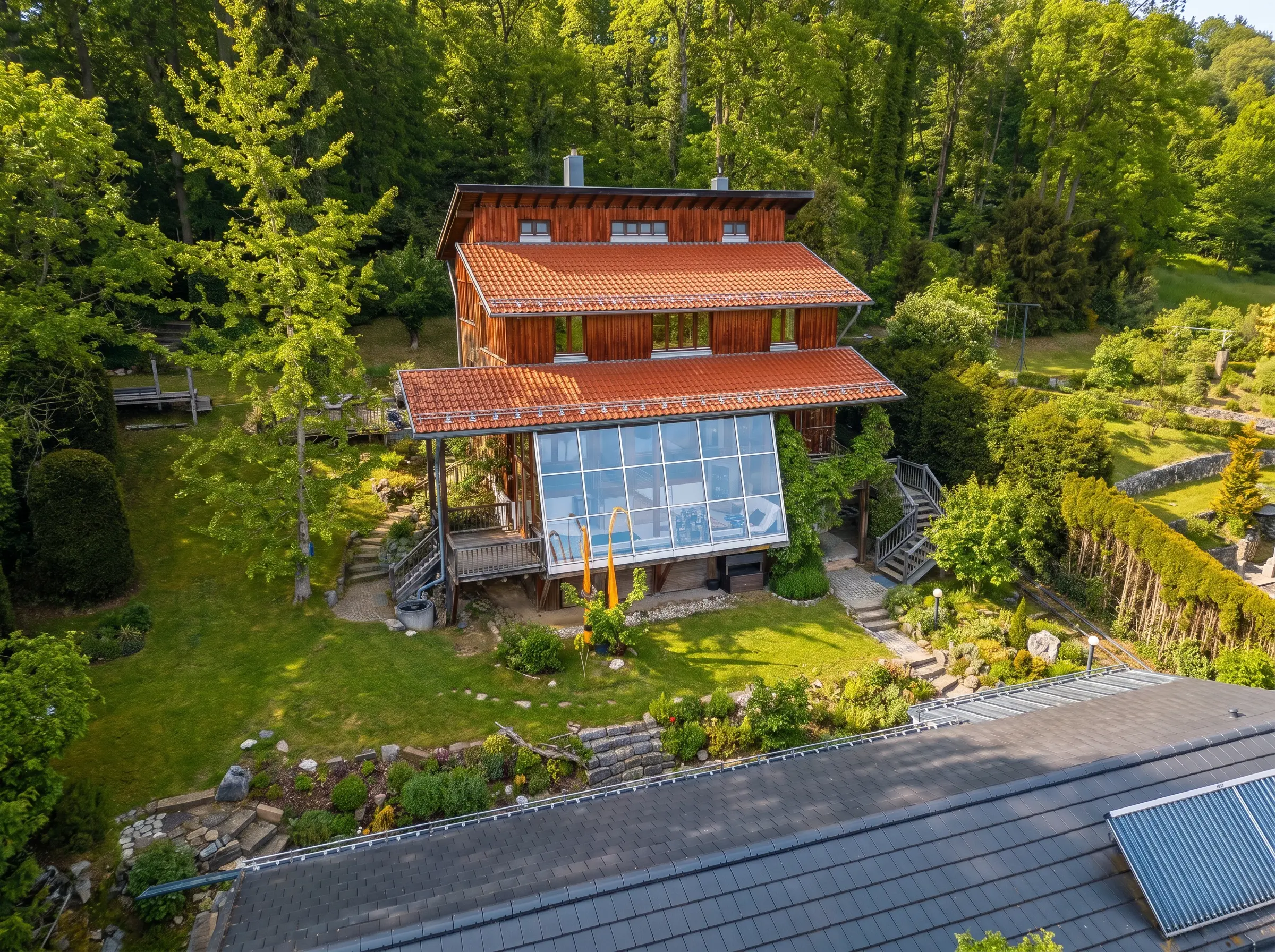 Ein Holzhaus mit rotem Ziegeldach und einer großen, verglasten Terrasse liegt inmitten üppiger Vegetation und gepflegter Gärten in einer waldreichen Gegend.