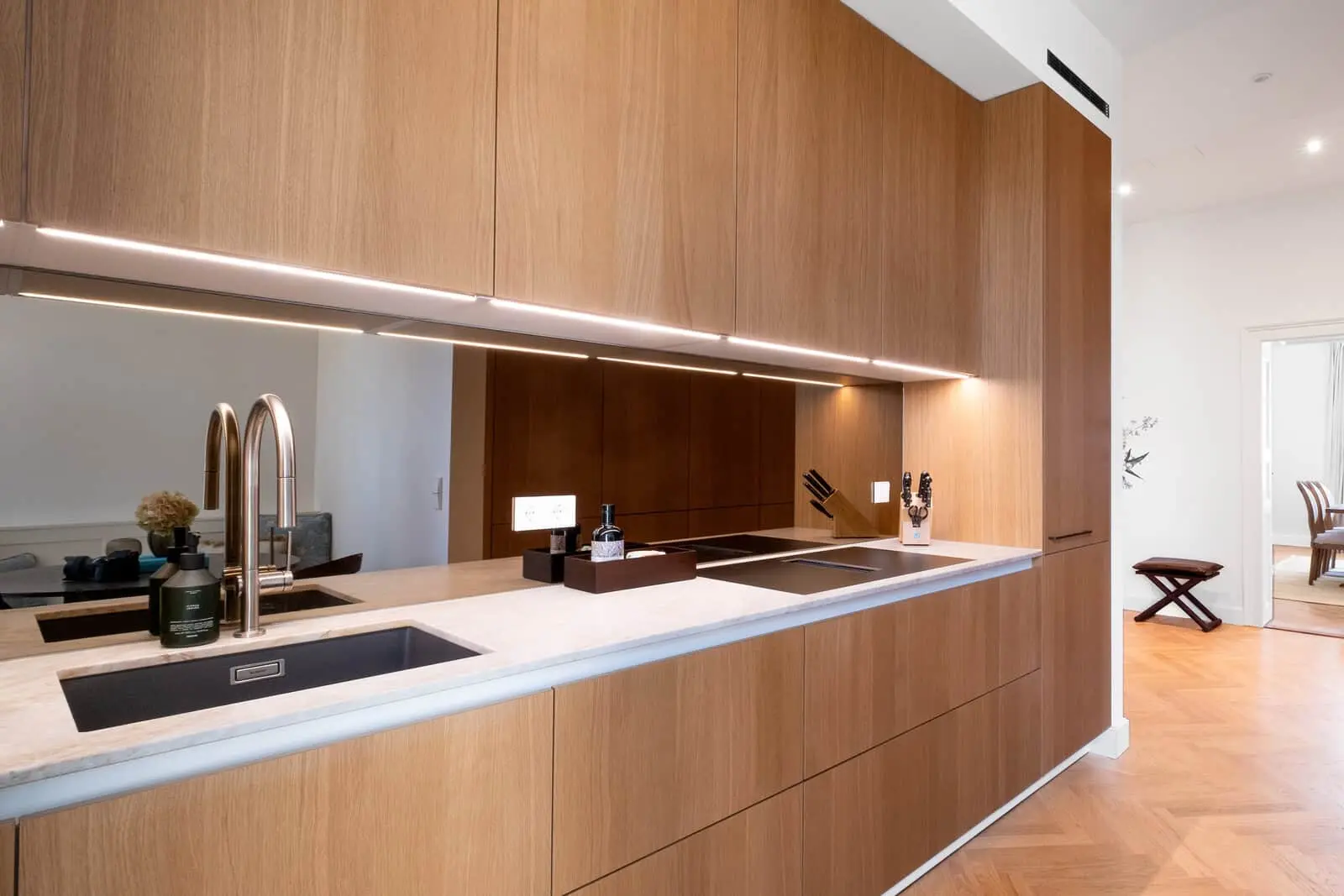 Moderne Küche mit Holzschränken, einer Arbeitsplatte aus Marmor, zwei Spülbecken mit Metallarmaturen, Unterschrankbeleuchtung und einer verspiegelten Aufkantung. Der Essbereich ist durch eine offene Tür sichtbar.