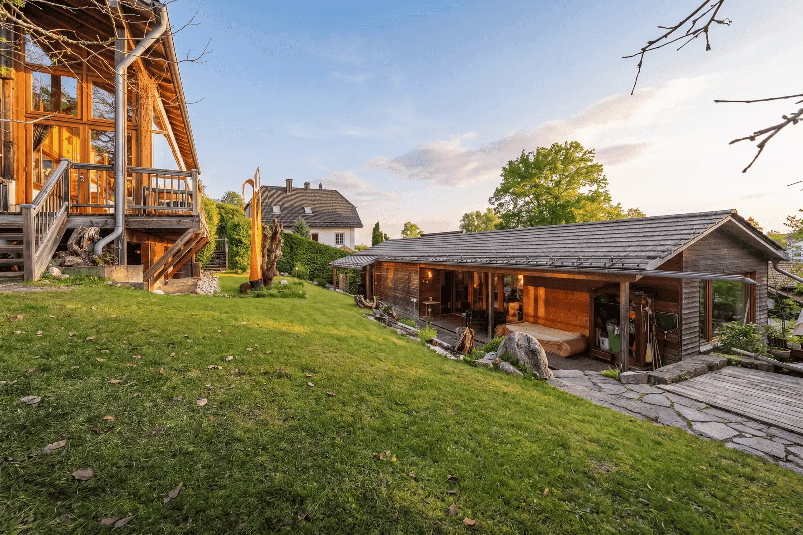 Ein modernes Holzhaus mit einer überdachten Veranda steht auf einem grasbewachsenen Rasen, im Hintergrund sind ein weiteres Haus und Bäume zu sehen, und der Himmel ist teilweise bewölkt.
