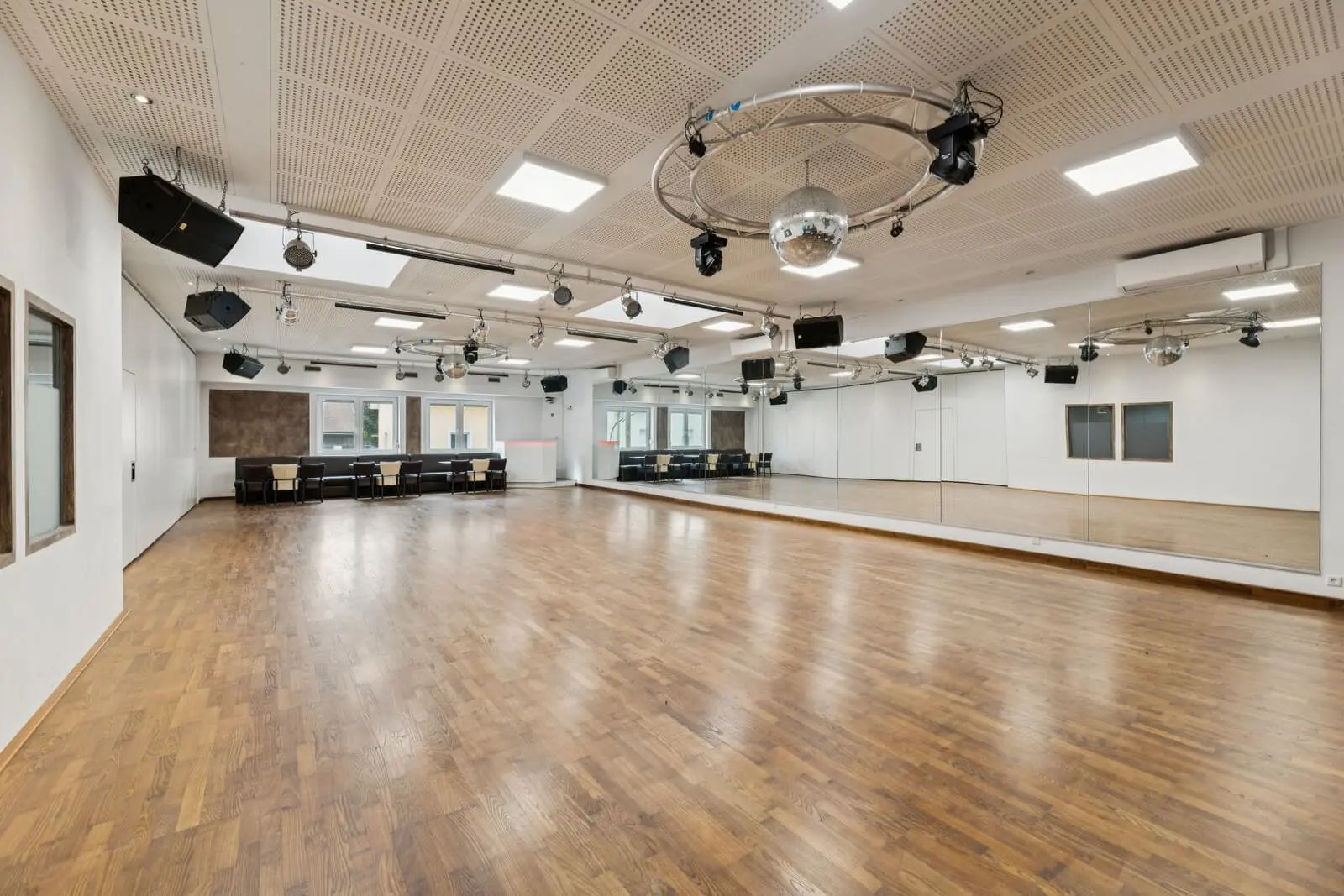 Geräumiges leeres Tanzstudio mit Holzboden, großen Spiegeln an einer Wand, Fenstern, Deckenbeleuchtung, Lautsprechern und einer Discokugel.