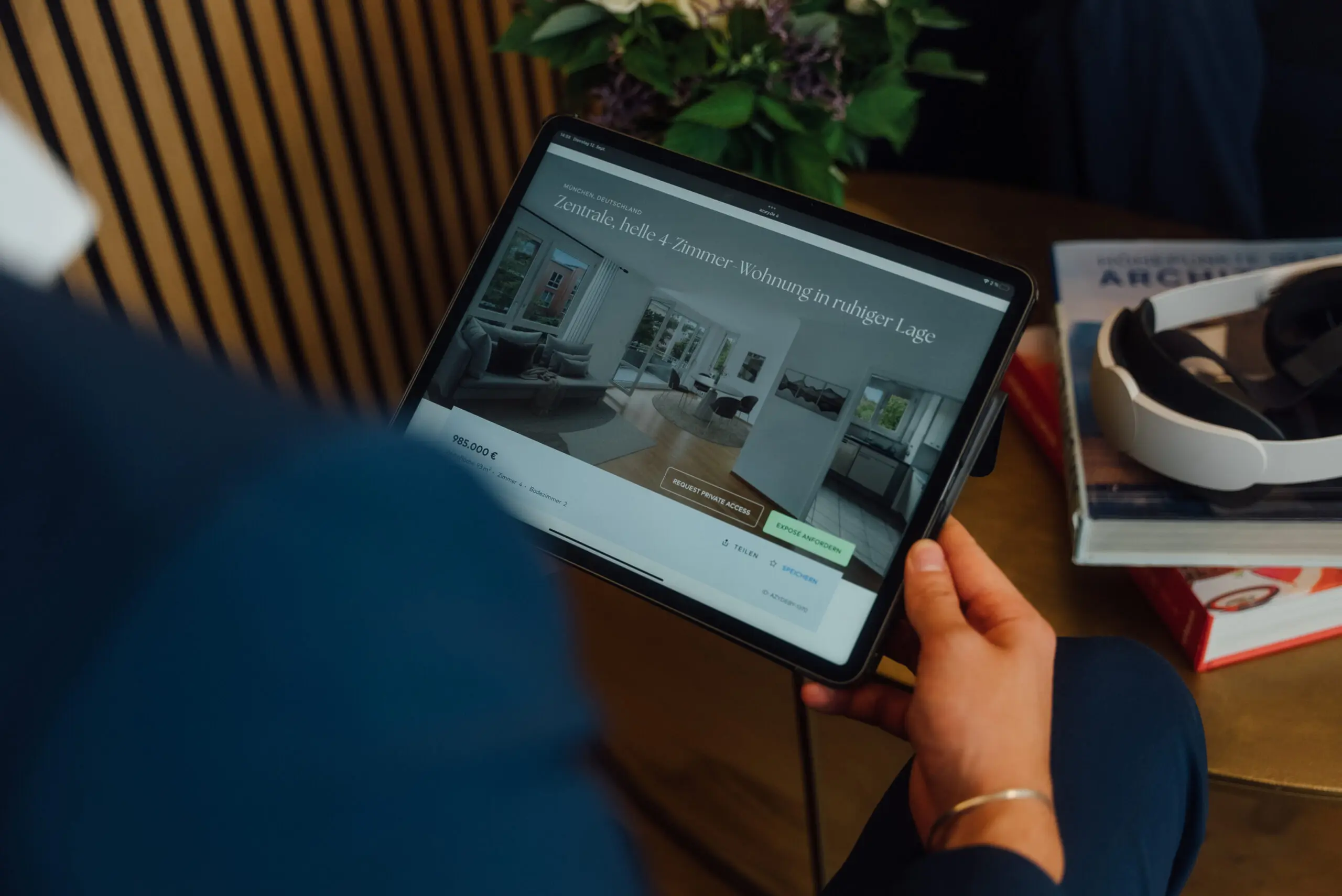 Person hält ein Tablet, auf dem eine Website mit Immobilienangeboten angezeigt wird, mit einem Virtual-Reality-Headset und Büchern auf einem Tisch im Hintergrund.