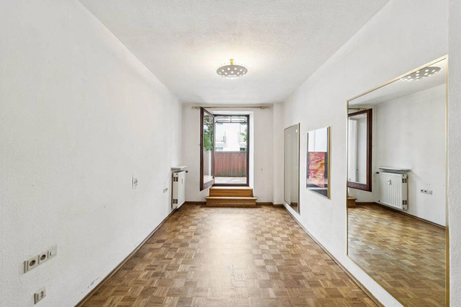 Leeres Zimmer mit weißen Wänden, Parkettboden, einem großen Wandspiegel, einem offenen Fenster mit Blick nach draußen und einem Heizkörper unter dem Fenster.