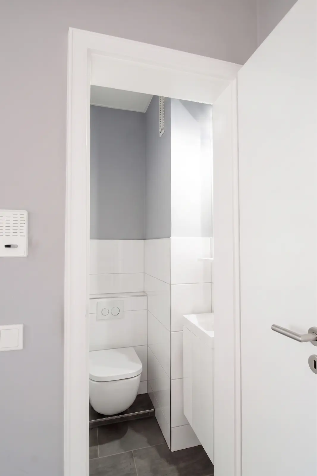Kleines, modernes Badezimmer mit weißen Fliesen, einem wandhängenden WC, einem kompakten Waschbecken und einer teilweise geöffneten Tür. Der Raum hat hellgraue und weiße Wände sowie dunkle Bodenfliesen.