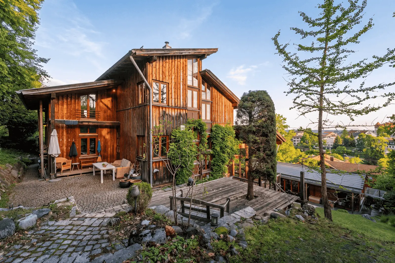 Ein großes Holzhaus mit mehreren Terrassen und Sitzgelegenheiten im Freien, umgeben von Bäumen und Grün an einem sonnigen Tag.