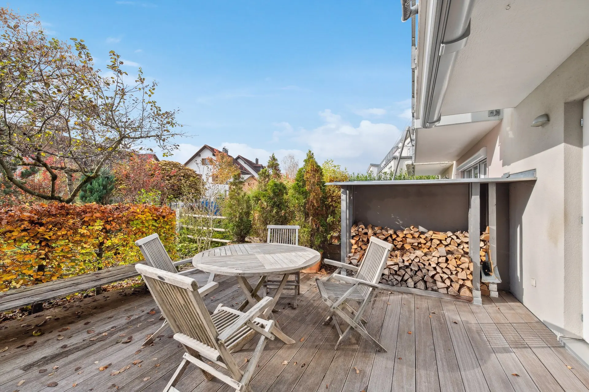 Holzterrasse mit rundem Tisch und Stühlen, umgeben von Bäumen und Sträuchern. Gestapeltes Brennholz wird unter einem Überhang gelagert. Unter einem klaren blauen Himmel sind nahe gelegene Häuser zu sehen.