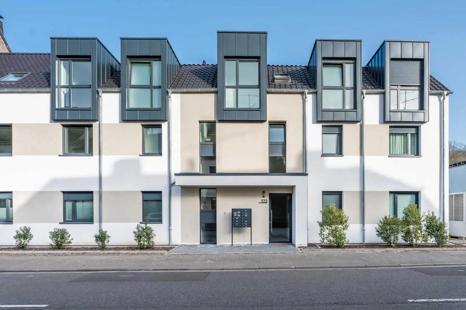 Modernes zweistöckiges Mehrfamilienhaus mit großen kastenförmigen Fenstern im Obergeschoss, neutraler Fassade und kleinen Sträuchern vor dem Haus entlang der Straße.