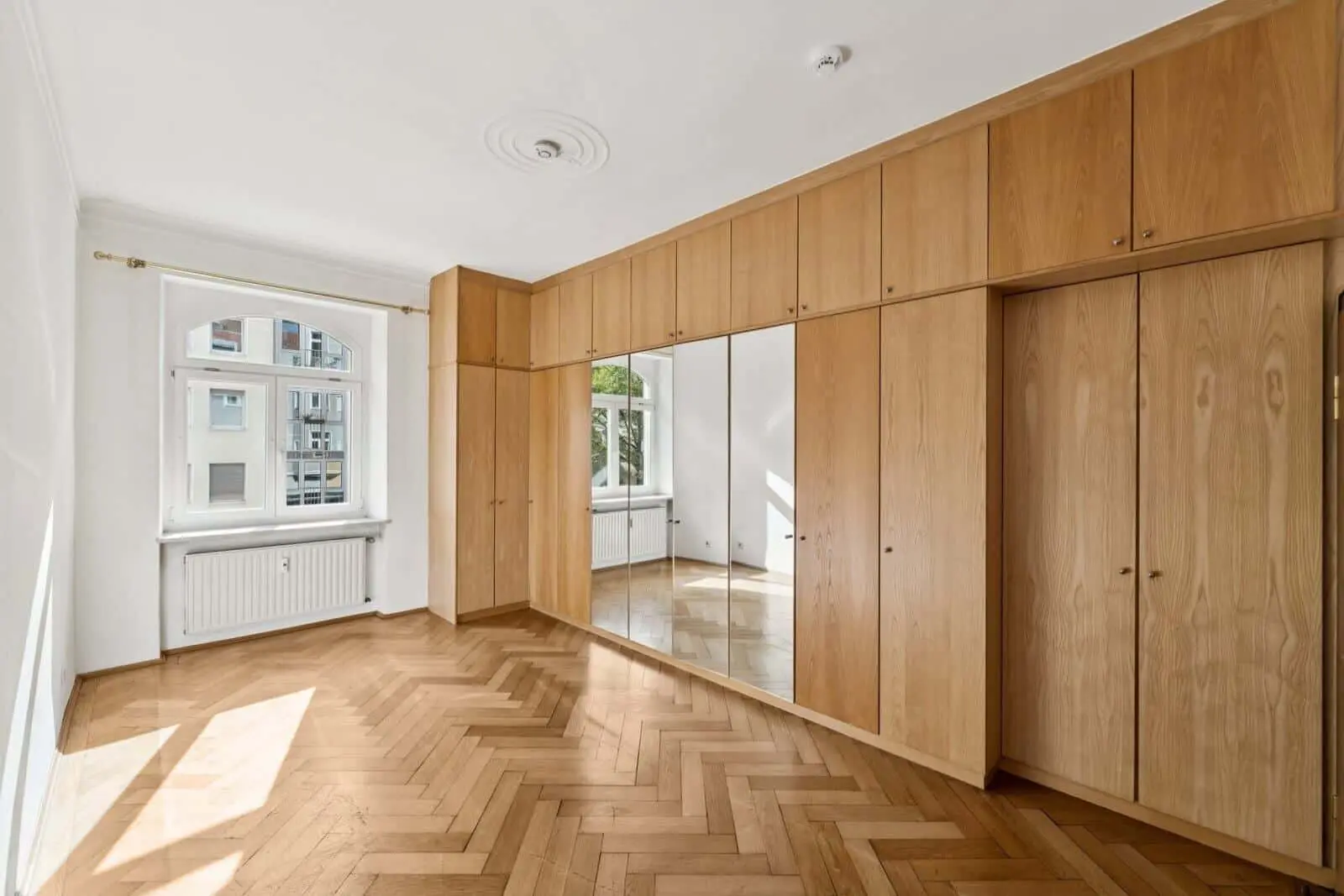 Helles, leeres Zimmer mit Fischgrätparkett, Holzeinbauschränken mit Spiegeln, großem Fenster und weißen Wänden.